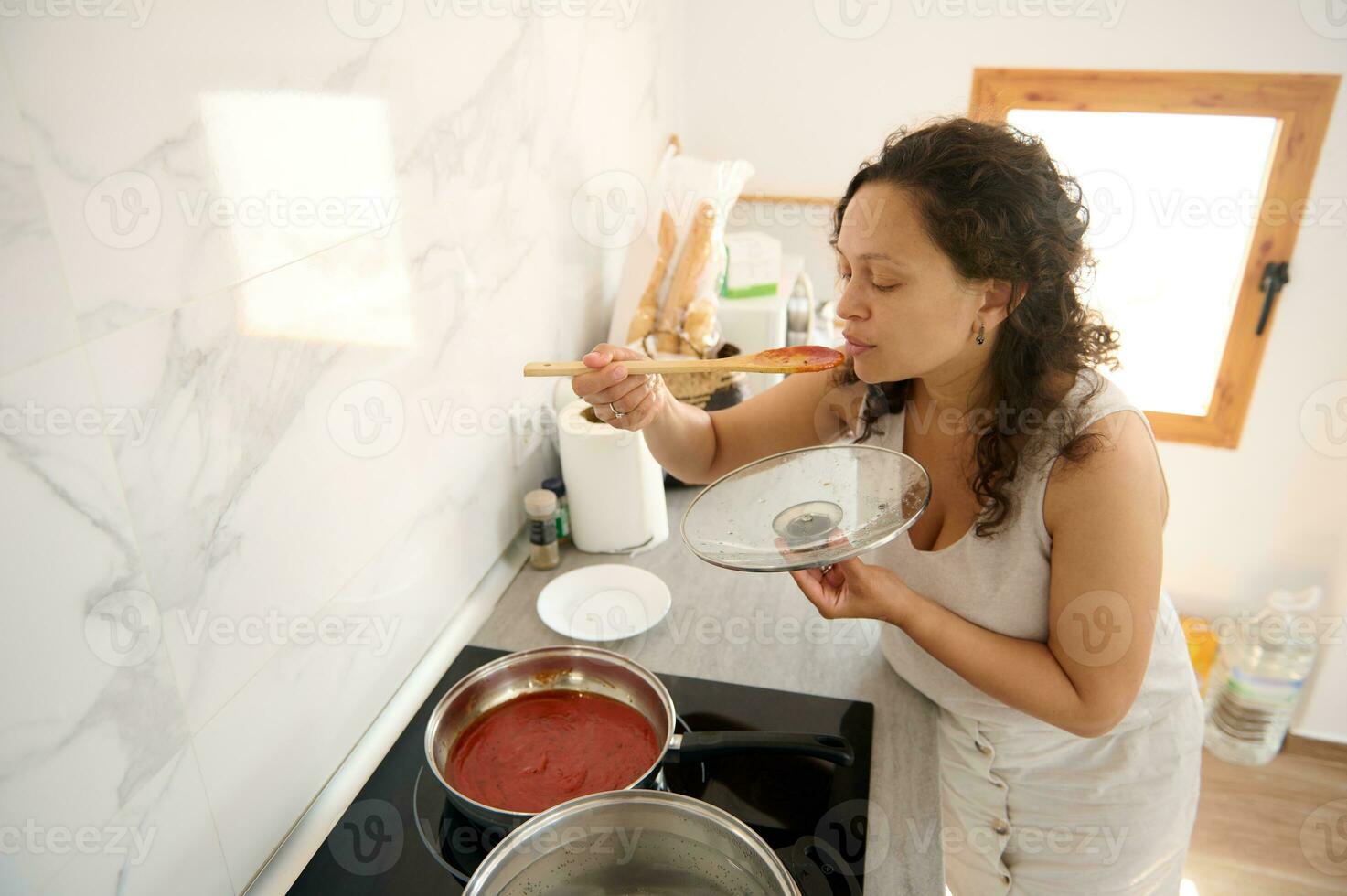 jong Latijns Amerikaans mooi huisvrouw proeverij de tomaat saus zij is Koken in de huis keuken foto