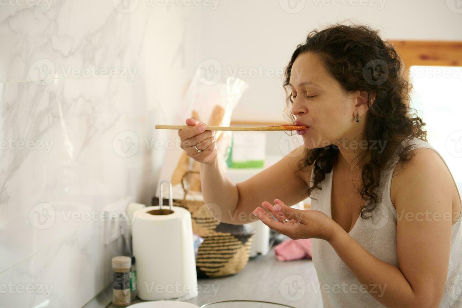 detailopname van een mooi vrouw huisvrouw proeverij een vers gekookt tomaat saus terwijl Koken avondeten in de huis keuken foto