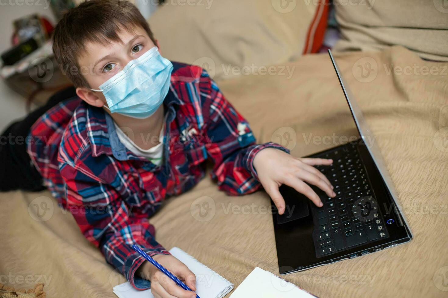 jongen in beschermend medisch masker leugens in de buurt laptop en schrijft in werk boek. kind doet lessen aan het kijken online uitzending lessen Aan internetten. e-learning, afgelegen aan het studeren internet concept foto