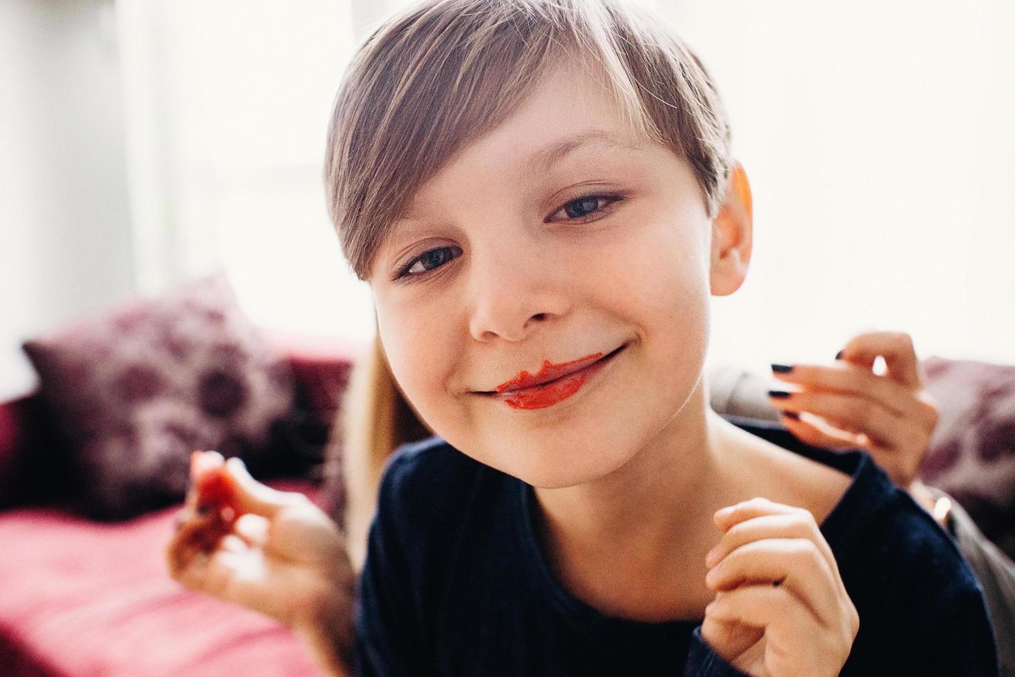 een zacht gefocust close-upbeeld van een schattige jongen met een gezicht beschilderd met eetbare felle kleurencrème, glimlachend met clownlippen, een lichte kamer op de achtergrond foto