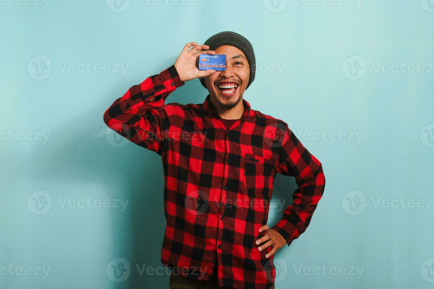 blij jong Aziatisch Mens met een muts hoed en rood plaid flanel overhemd is speels aan het bedekken een oog met een credit kaart terwijl staand tegen een blauw achtergrond foto