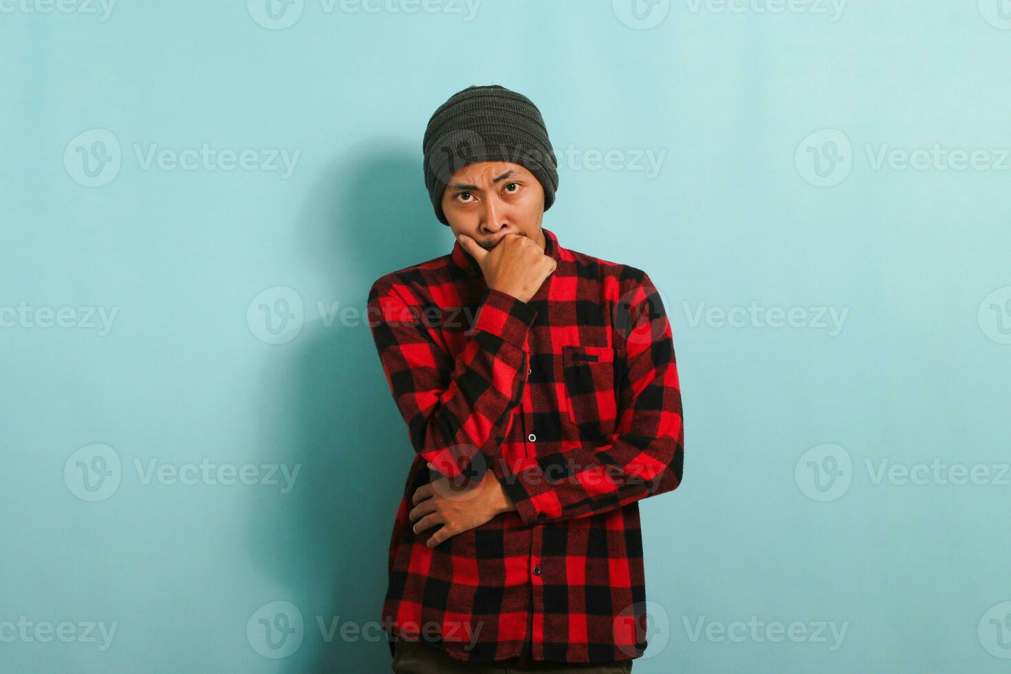 attent jong Aziatisch Mens met muts hoed en rood plaid flanel overhemd houdt zijn kin en looks peinzend Bij camera, maken plannen, geïsoleerd Aan een blauw achtergrond foto