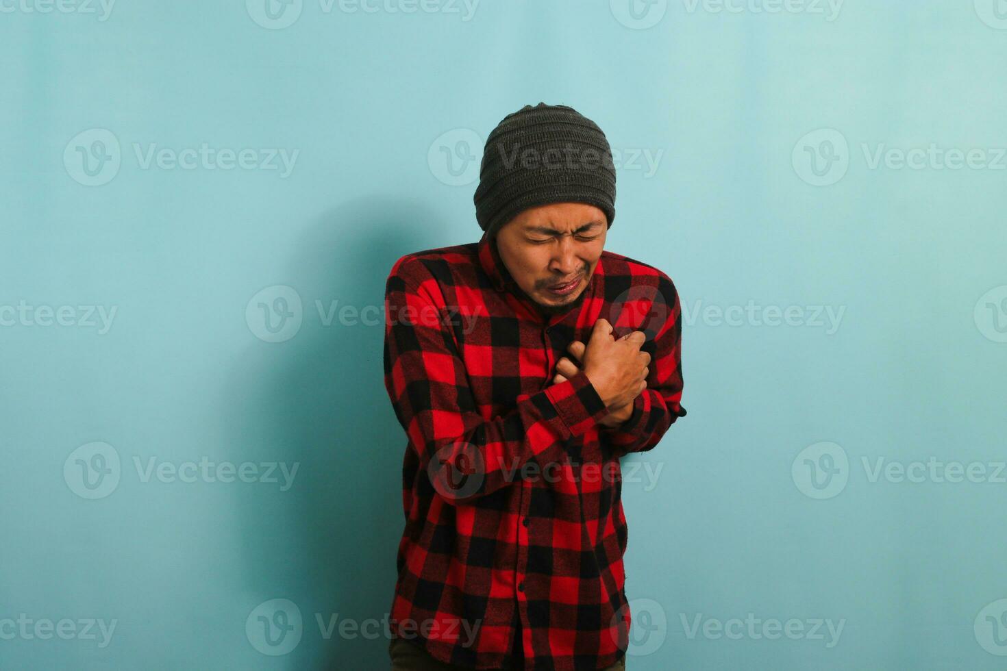 een jong Aziatisch Mens met een muts hoed en een rood plaid flanel overhemd koppelingen zijn borst, ervaren acuut pijn, welke mei worden een symptoom van een hart aanval, terwijl staand tegen een blauw achtergrond foto