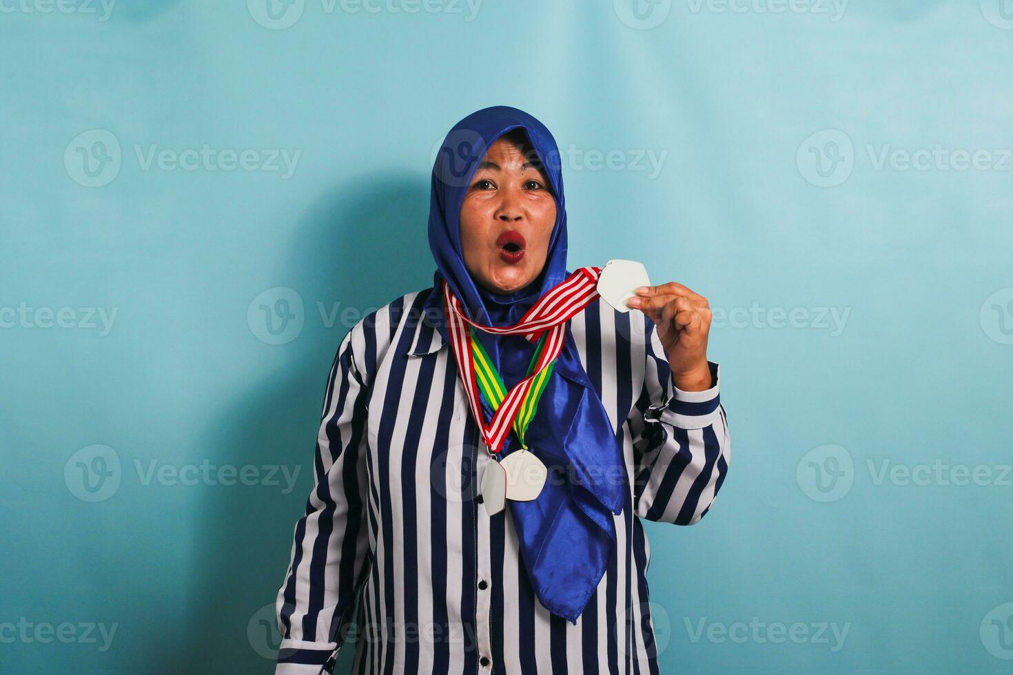 een opgewonden middelbare leeftijd Aziatisch vrouw in een blauw hijab en een gestreept overhemd is presentatie van een medaille terwijl wezen geïsoleerd Aan een blauw achtergrond foto