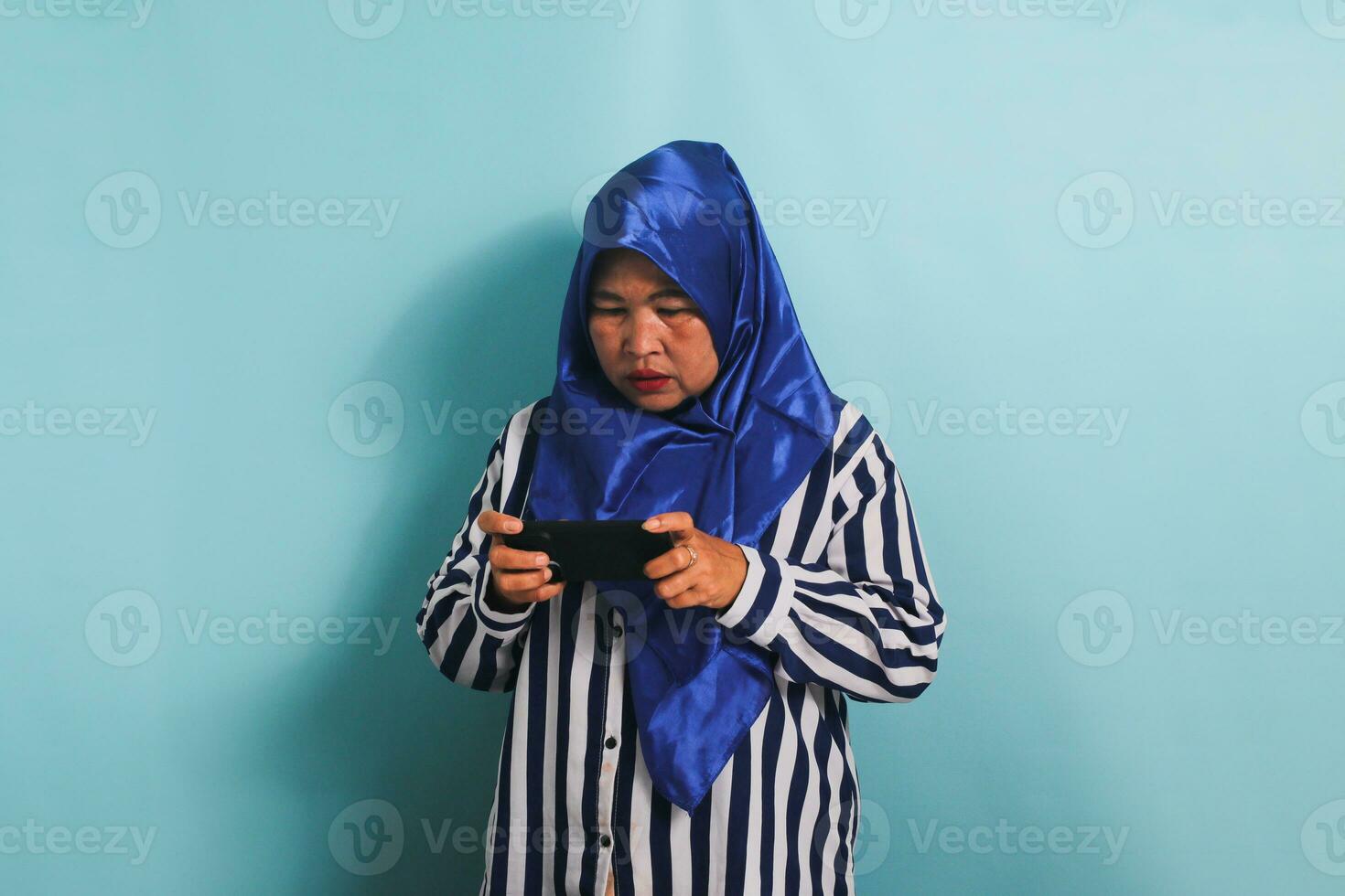 een verveeld middelbare leeftijd Aziatisch vrouw in een blauw hijab en een gestreept overhemd is spelen een spel Aan haar mobiel telefoon, gebaren aarzelend, op zoek verdrietig, en verliezende de spel, geïsoleerd Aan een blauw achtergrond. foto