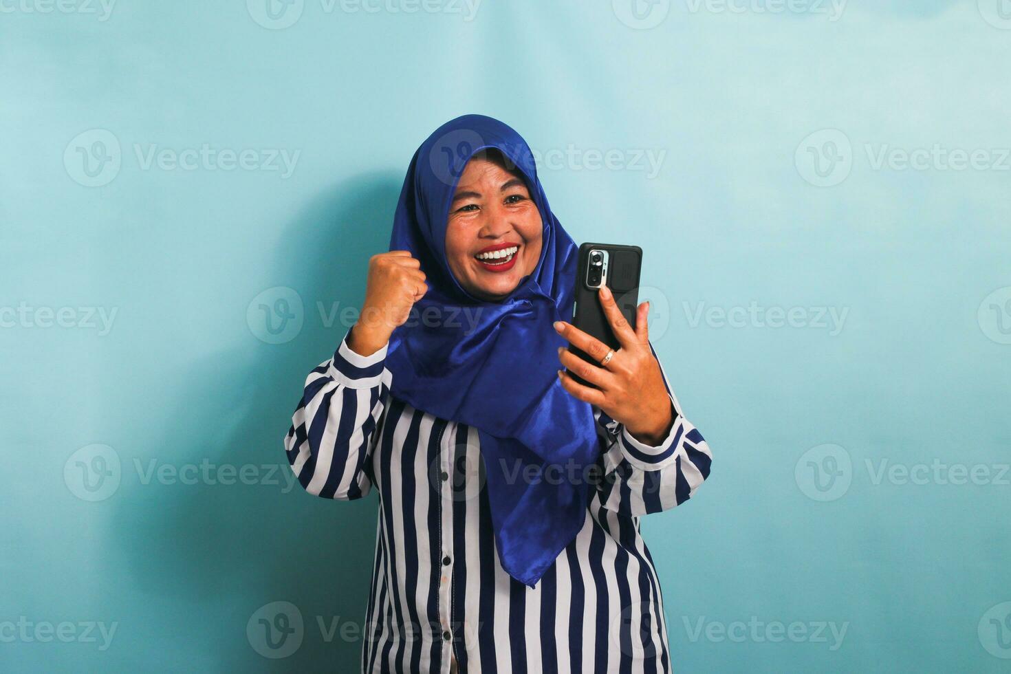 een opgewonden middelbare leeftijd Aziatisch vrouw in een blauw hijab en een gestreept overhemd is tonen een enthousiast uitdrukking terwijl Holding een mobiel telefoon. ze is geïsoleerd Aan een blauw achtergrond foto