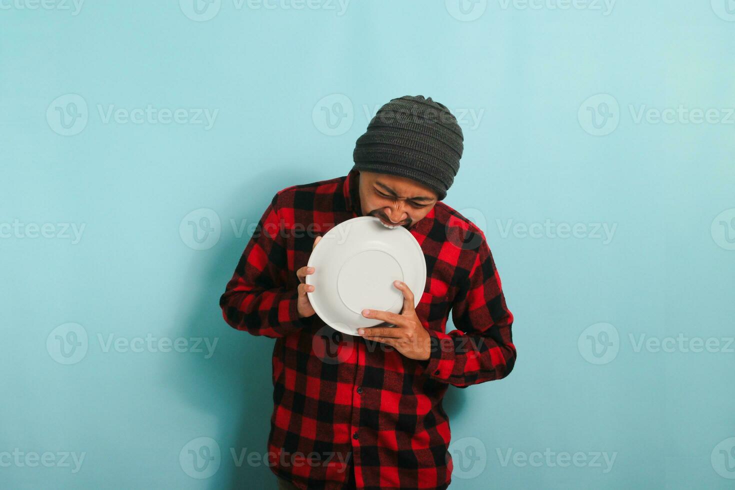 hongerig jong Aziatisch Mens bijt een leeg wit bord, geïsoleerd Aan een blauw achtergrond foto