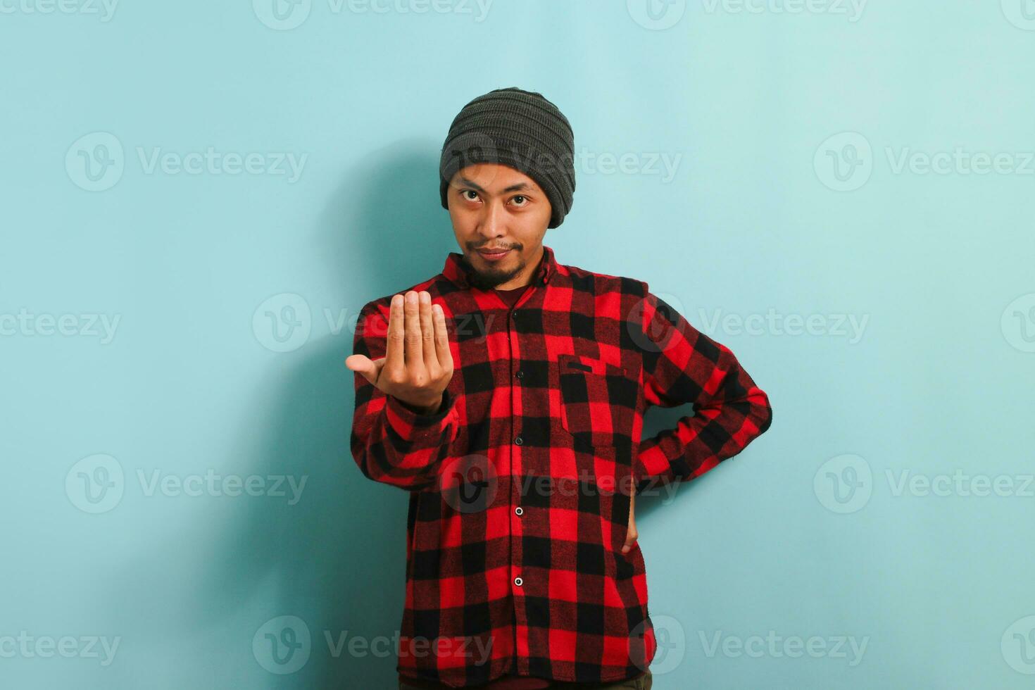 een jong Aziatisch Mens vervelend een muts hoed en een rood plaid flanel overhemd is maken een komen hier gebaar met zijn hand, uitnodigend, gastvrij, Vrolijk, en lachend. hij is geïsoleerd Aan een blauw achtergrond foto