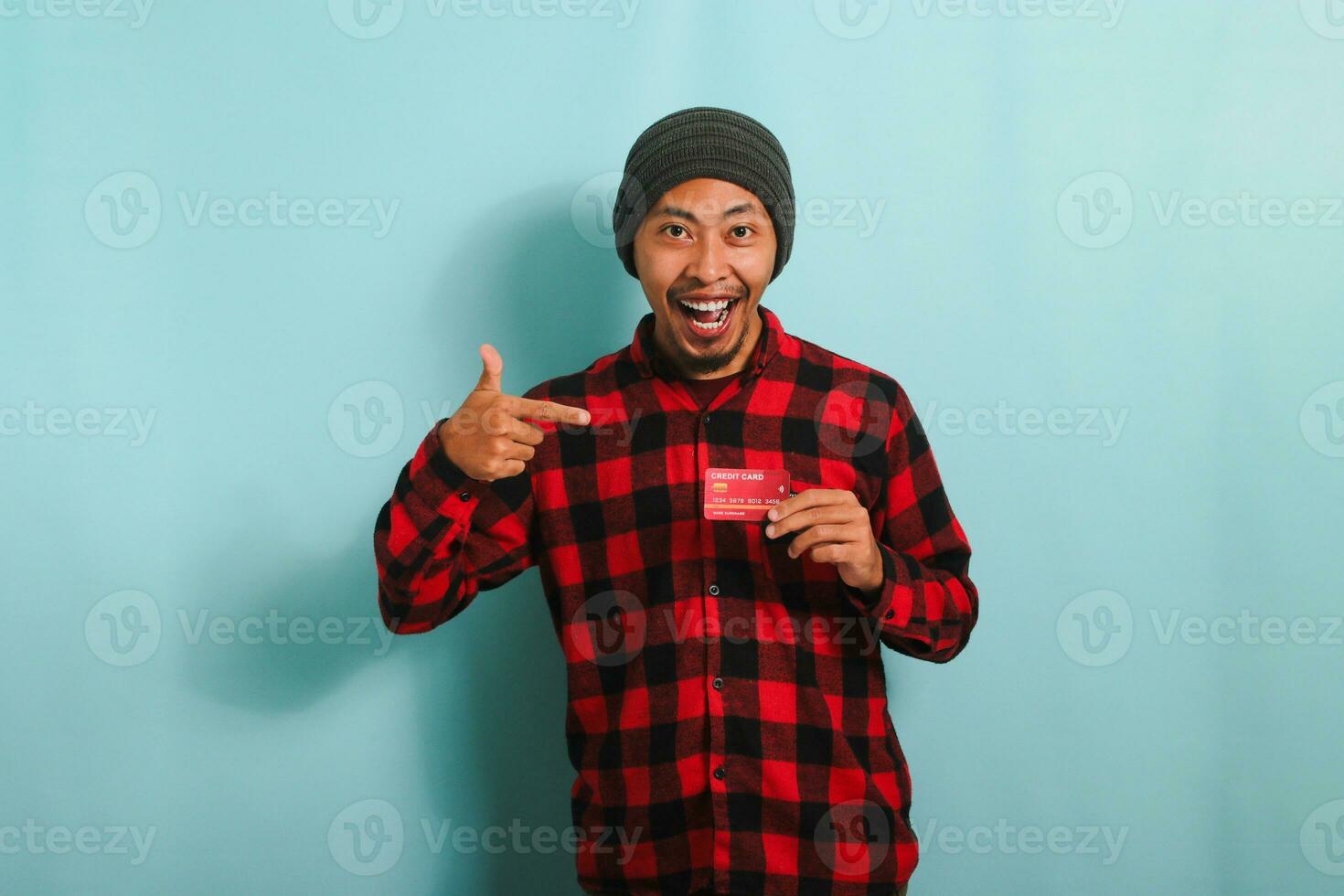 opgewonden jong Aziatisch Mens met een muts hoed en rood plaid flanel overhemd is richten naar zijn bank credit kaarten in zijn handen terwijl op zoek Bij camera en staand tegen een blauw achtergrond foto