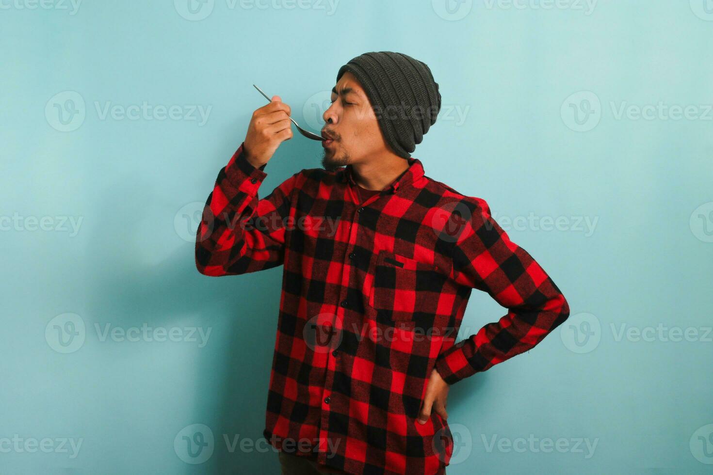 jong Aziatisch Mens met muts hoed en rood plaid flanel overhemd sluitend zijn ogen terwijl proeverij voedsel met een lepel, geïsoleerd Aan een blauw achtergrond foto