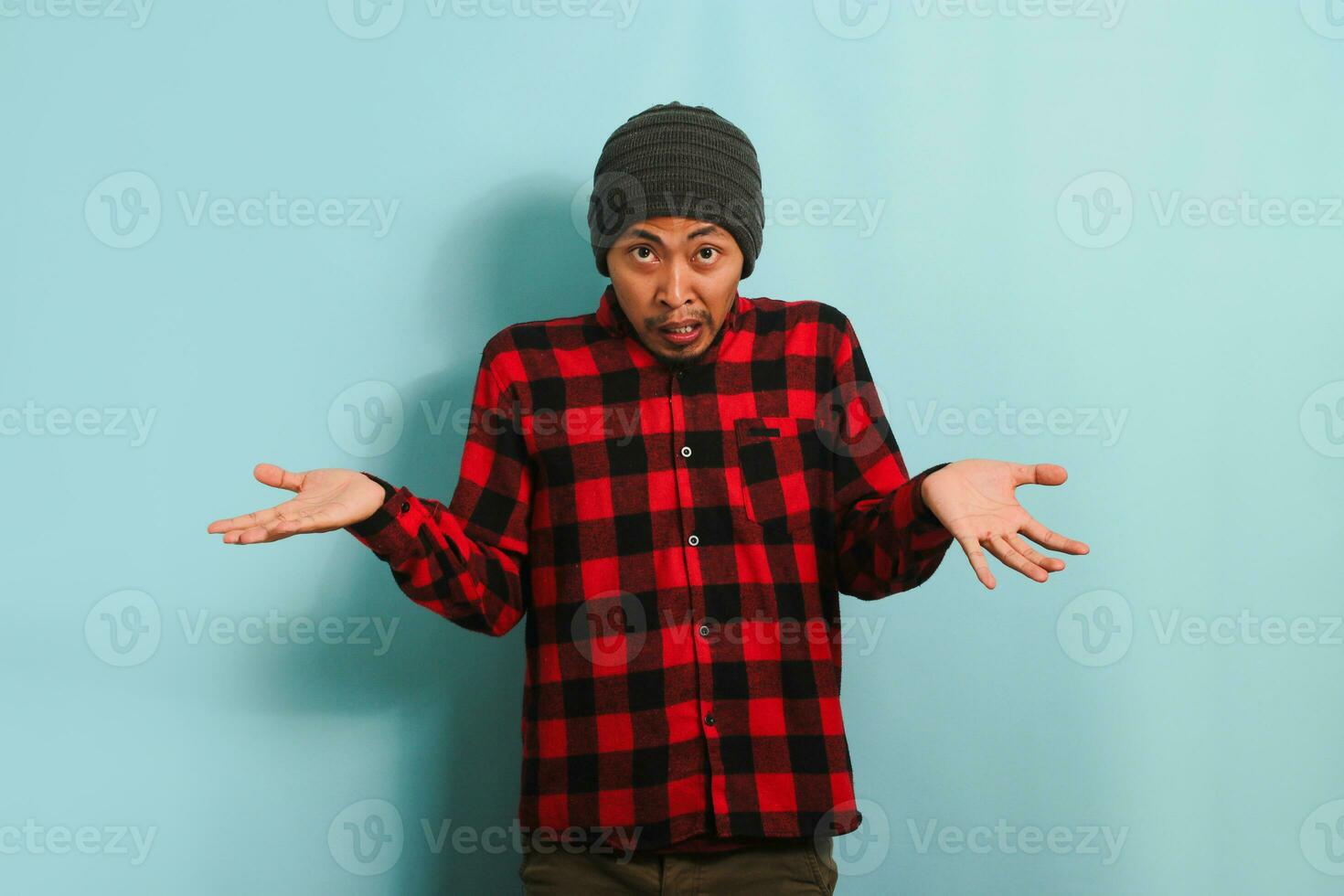 een verward jong Aziatisch Mens met een muts hoed en een rood plaid flanel overhemd is schouderophalend zijn schouders en op zoek Bij de camera met een verbaasd uitdrukking, geïsoleerd Aan een blauw achtergrond. foto