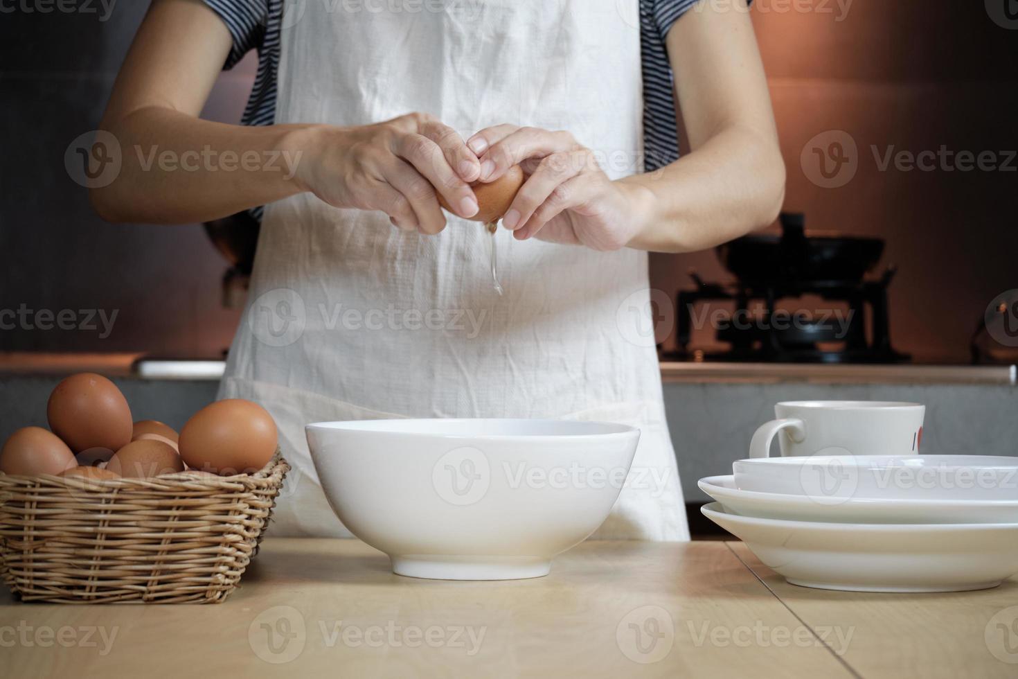 vrouwelijke kok in een witte schort kraakt een ei in de keuken van het huis. foto