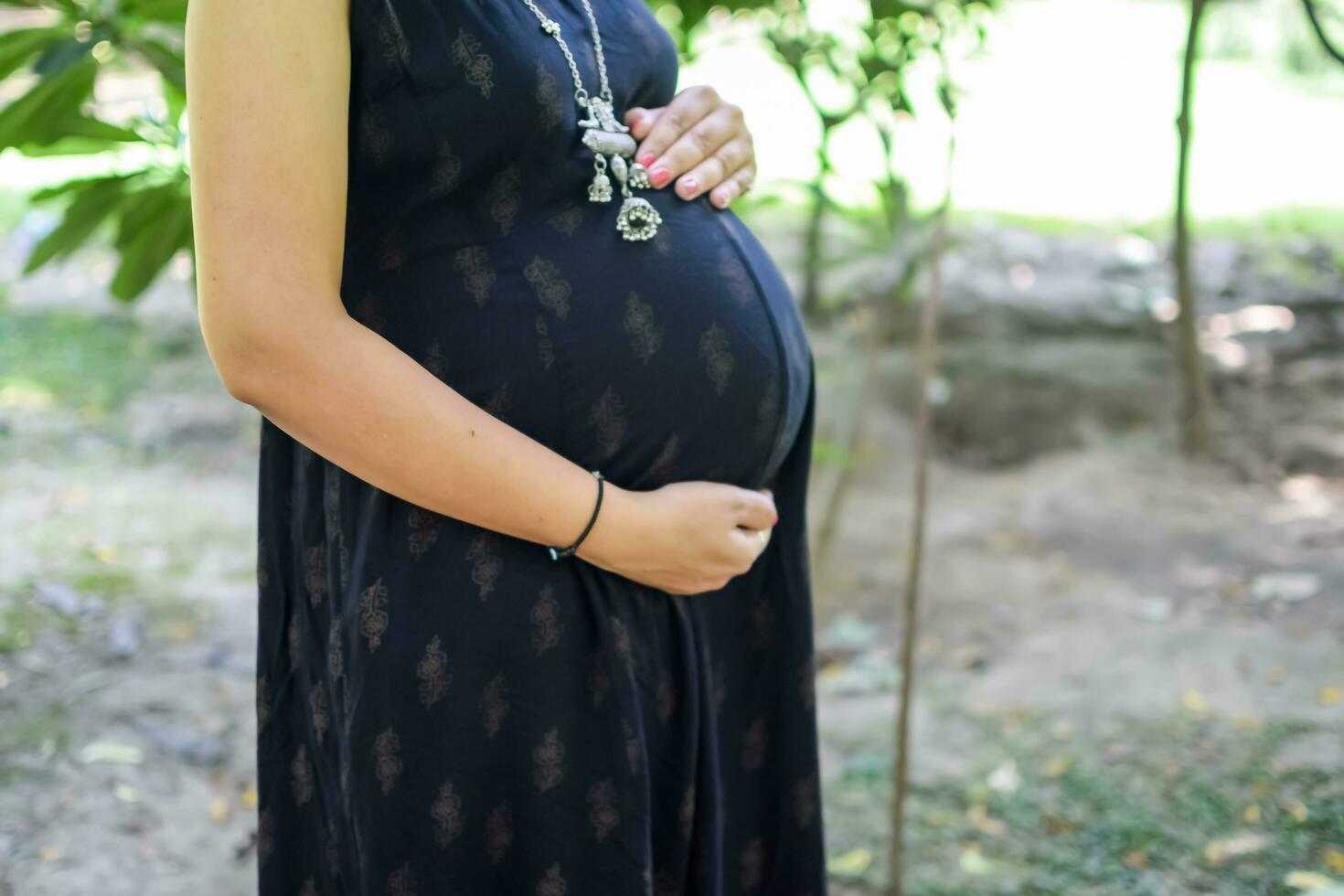 een zwanger Indisch dame poses voor buitenshuis zwangerschap schieten en handen Aan buik, Indisch zwanger vrouw zet haar hand- Aan haar maag met een moederschap jurk Bij maatschappij park, zwanger buiten moederschap schieten foto