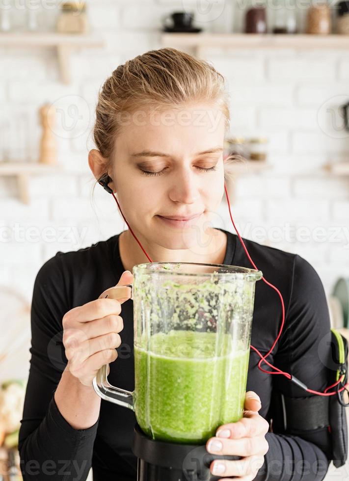 jonge blonde glimlachende vrouw die groene smoothie maakt thuis keuken foto