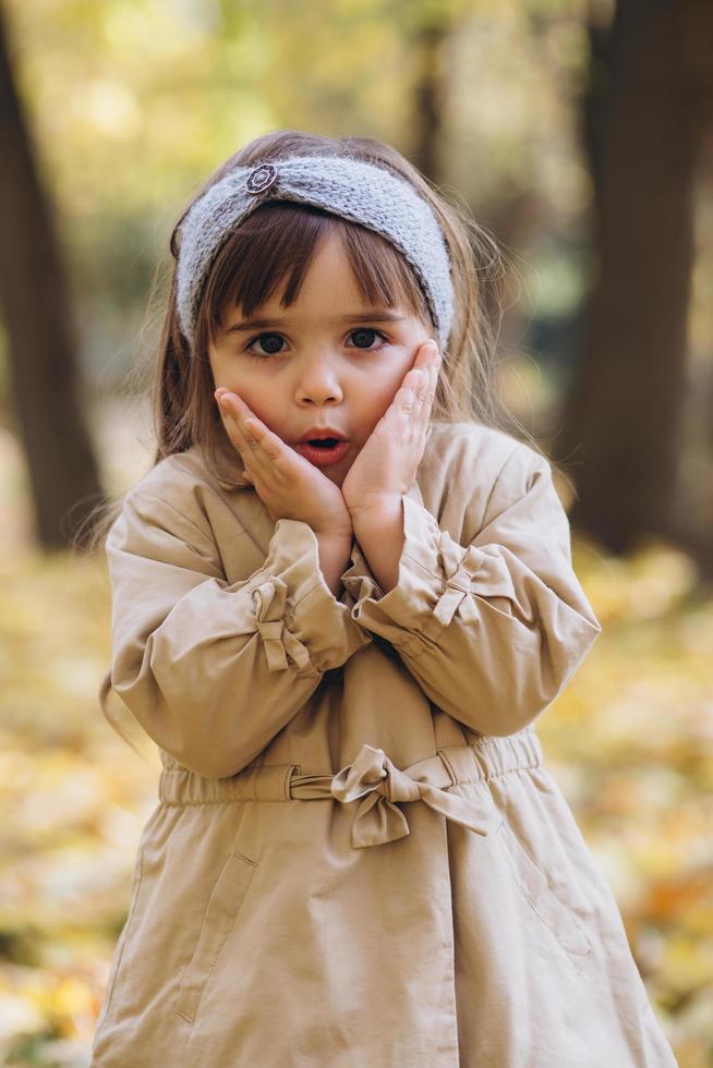 klein meisje in een beige jas toont emoties in het herfstpark foto