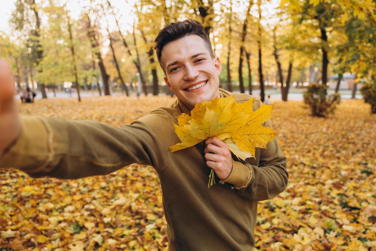 man die een boeket herfstbladeren vasthoudt en een selfie maakt in het park foto