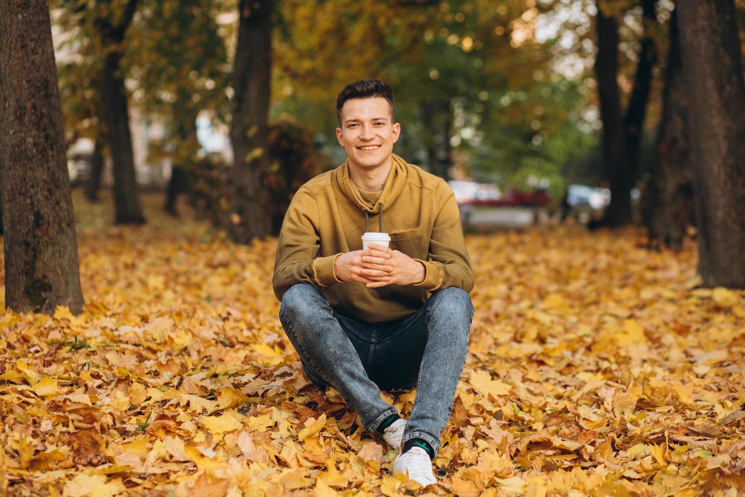 knappe en gelukkige jongen die lacht en koffie drinkt in het herfstpark foto