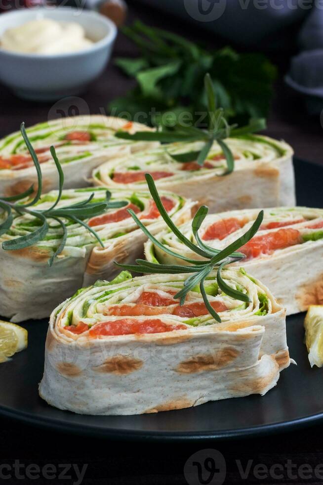 broodjes van dun pita brood en rood gezouten Zalm met sla bladeren Aan een zwart keramisch bord, donker houten achtergrond. foto