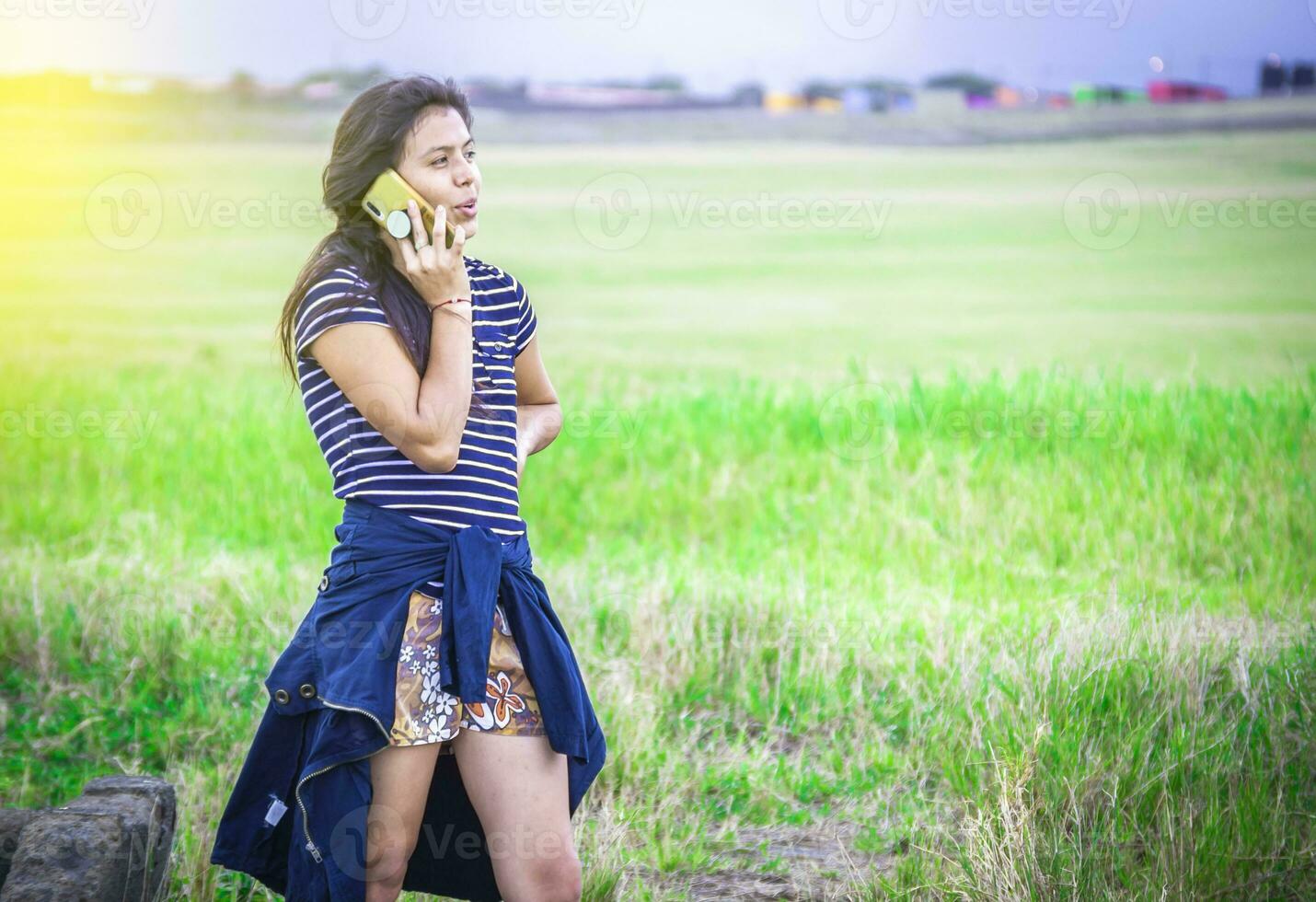 jong meisje roeping telefoon Aan de groen veld, mooi jong vrouw roeping door cel telefoon Aan de gras buitenshuis. foto