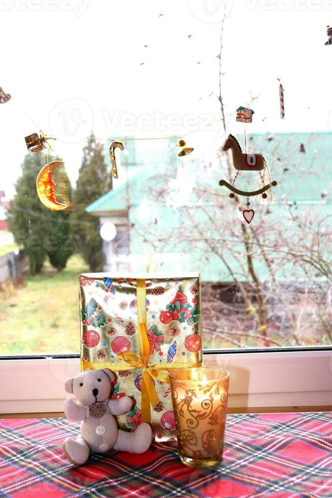 teddy beer met een Kerstmis geschenk en een kaars in een gouden glas kandelaar door de venster in een landelijk huis, verticaal foto