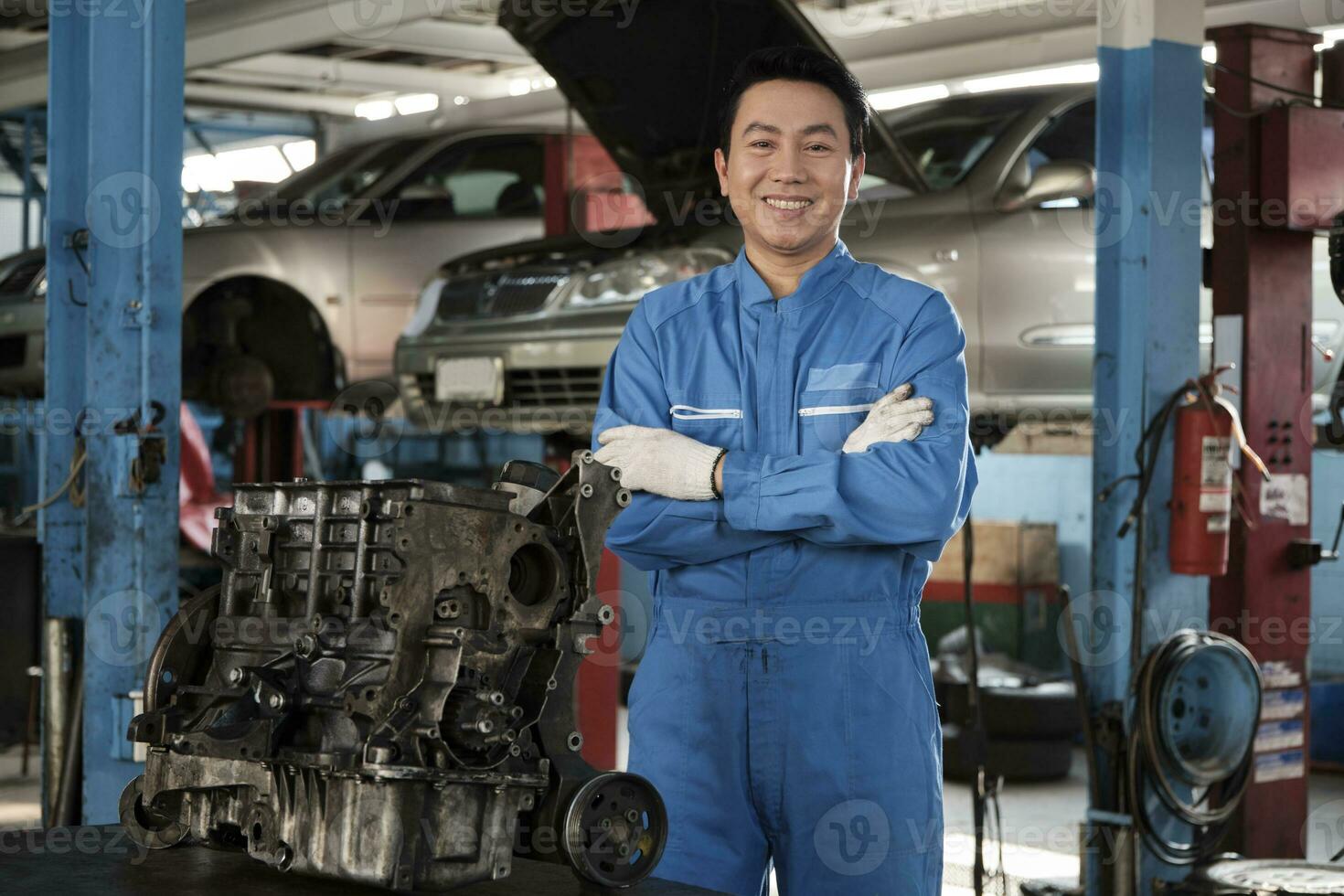 portret van Aziatisch mannetje monteur leidinggevende in uniform, armen gekruiste en op zoek Bij camera in de buurt auto motor Bij onderhoud garage, gelukkig onderhoud werk, controleren en reparatie bezetting in automotive industrie. foto