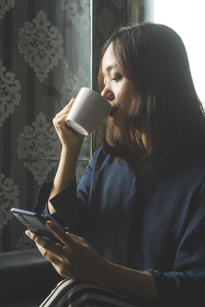 ontspannen aziatische vrouw die koffie drinkt terwijl ze een mobiele telefoon gebruikt voor werk foto