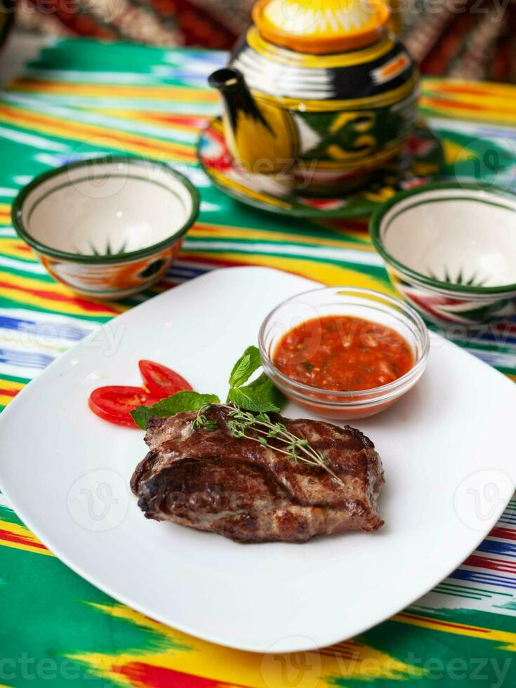 rundvlees steak met tomaat saus, tomaten en kruiden in oosters stijl Aan een tafel met een theepot en een kop voor thee. foto