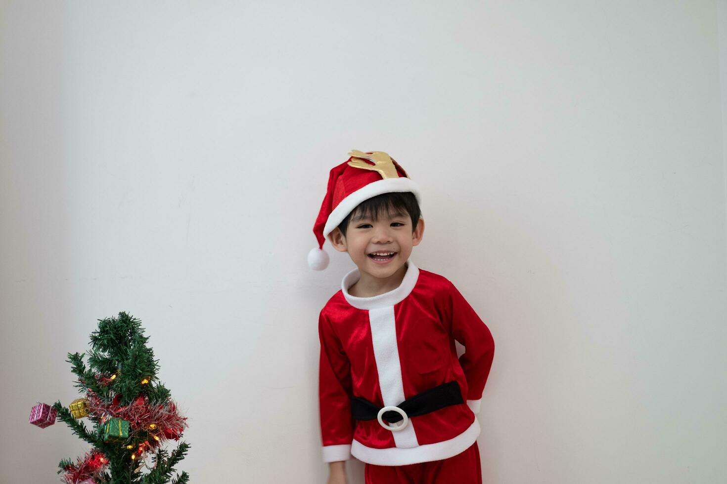 Aziatisch jongen vervelend de kerstman kostuum staand en spelen in de buurt een pret Kerstmis boom Aan wit achtergrond. foto