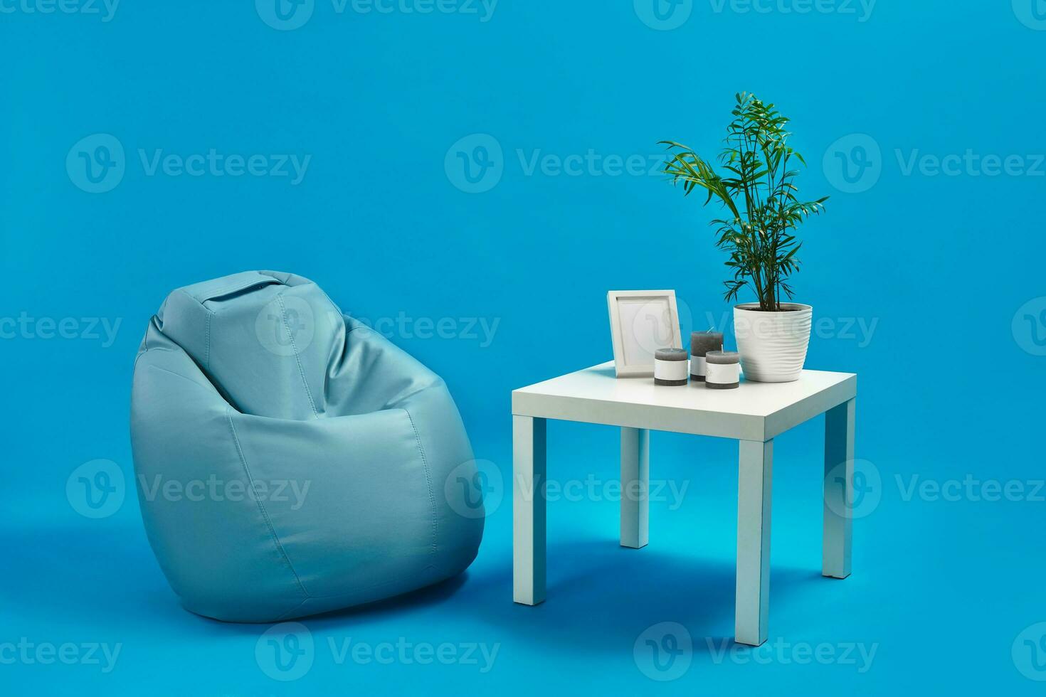 turkoois fauteuil Boon zak en wit koffie tafel met groen bloem in een pot, drie kaarsen en foto kader Aan het. blauw studio achtergrond. kopiëren ruimte