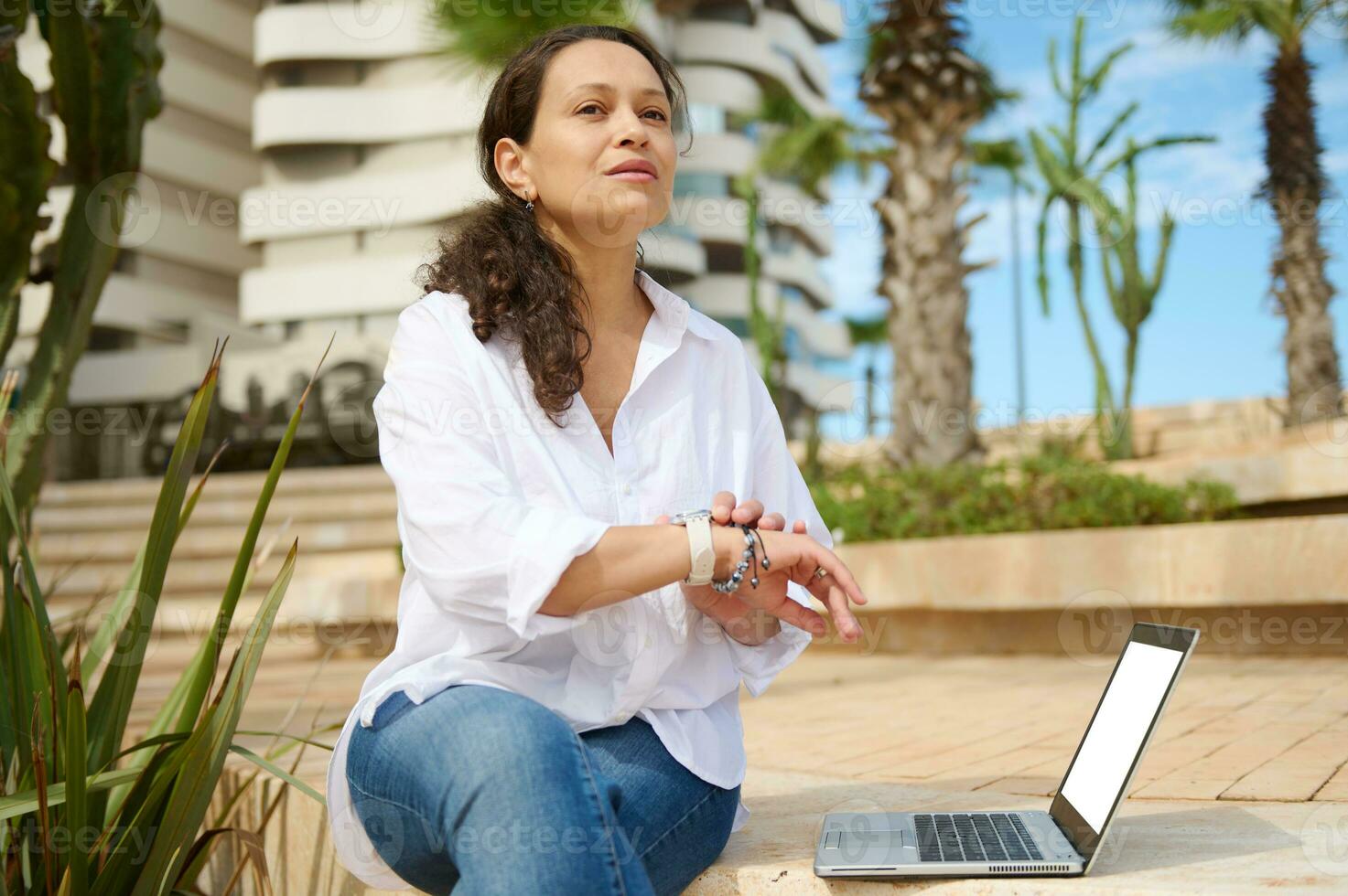 aantrekkelijk bedrijf vrouw freelancer in wit overhemd en gewoontjes jeans, controle haar slim horloge, peinzend op zoek terzijde foto