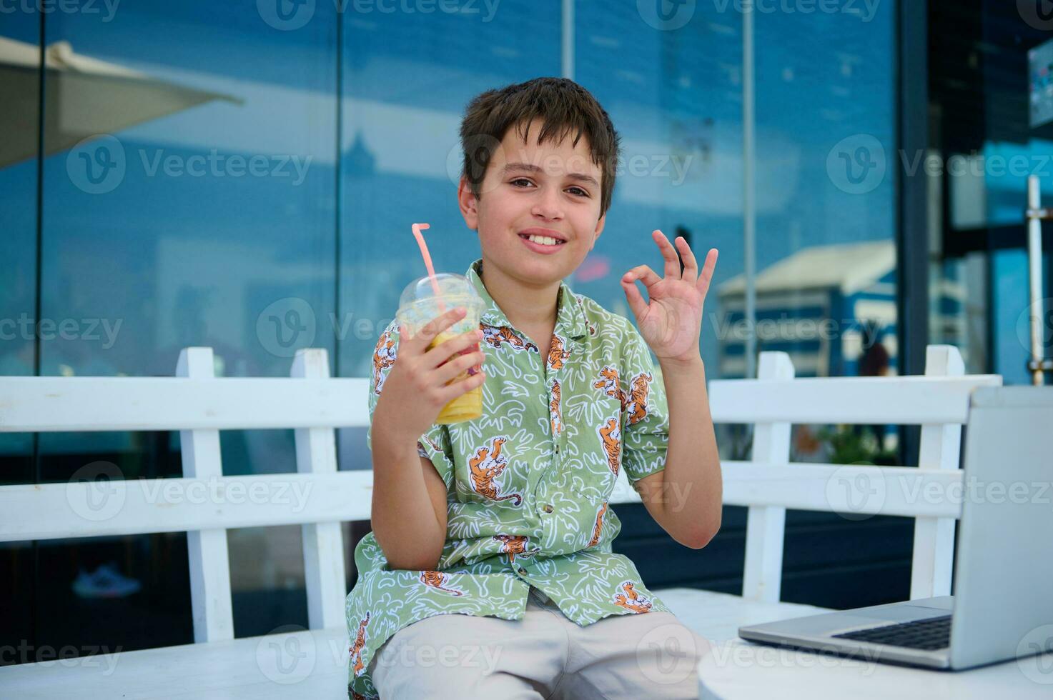 knap pre tiener jongen in zomer shirt, Holding een glas van fris geperst oranje sap, tonen OK hand- teken, vol vertrouwen op zoek Bij camera foto