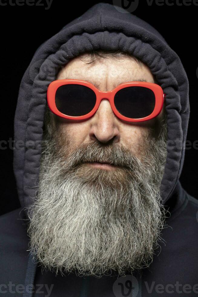 een ouderen Mens met een grijs baard in een kap en rood bril Aan een zwart achtergrond. een Mens verbergt iets. foto