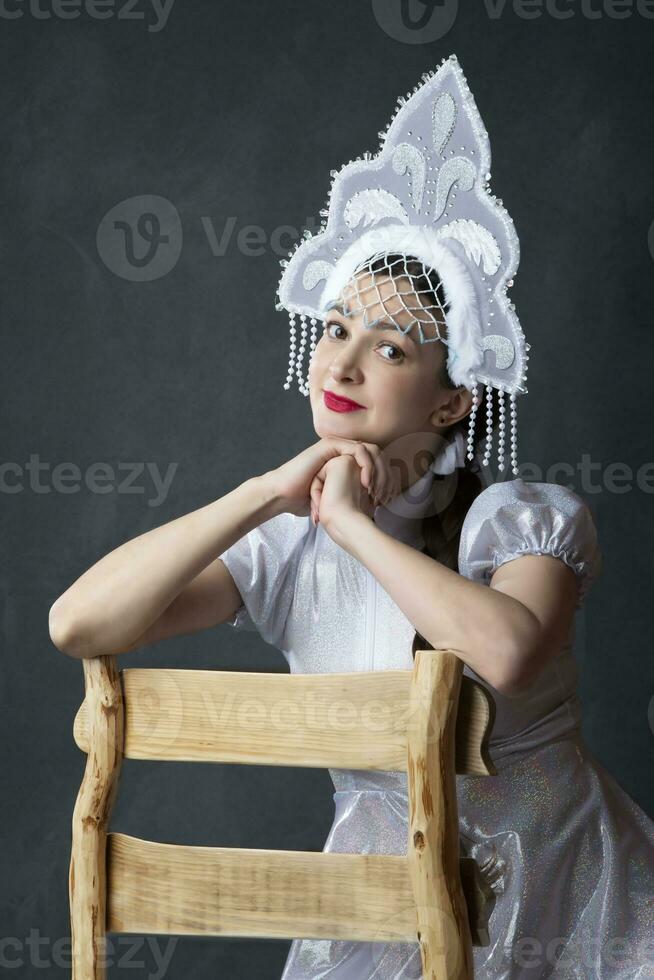 portret van een mooi meisje in een sneeuw meisje kostuum Aan een grijs achtergrond. een Russisch sneeuw meisje in een kroon zit Aan een houten stoel. foto