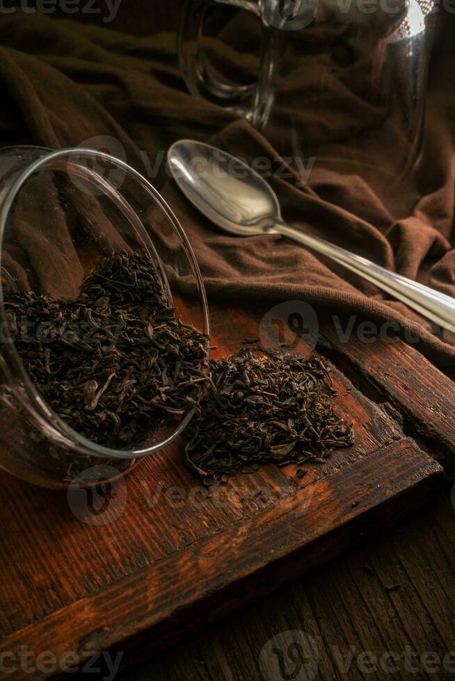 heerlijk droog los blad thee is gegoten uit van een glas pot Aan een houten wijnoogst bruin tafel met een lepel, een kleding en een glas Aan een donker achtergrond foto