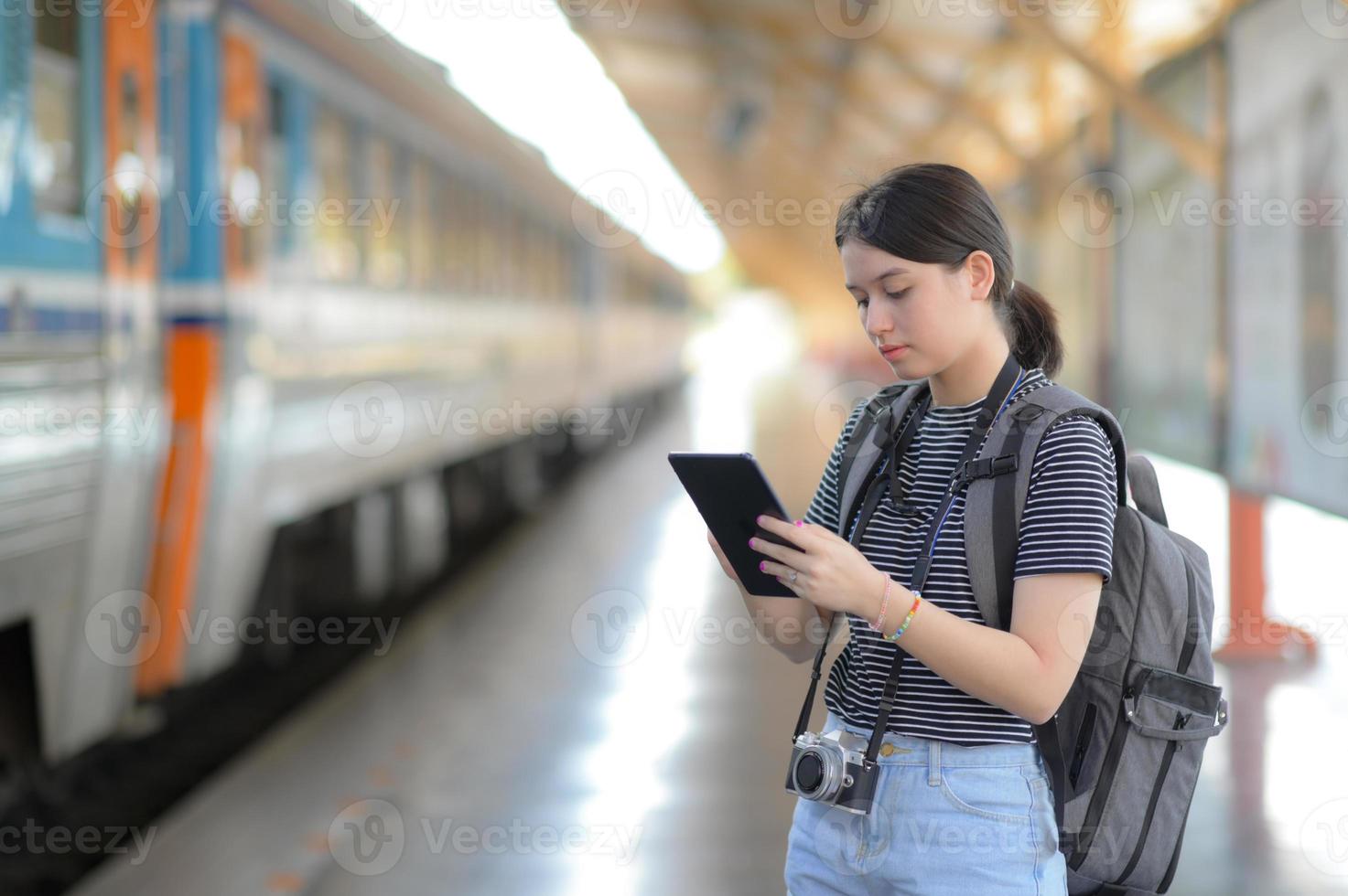 een buitenlandse vrouwelijke reiziger draagt een rugzak met behulp van een tablet terwijl ze op een trein wacht. foto
