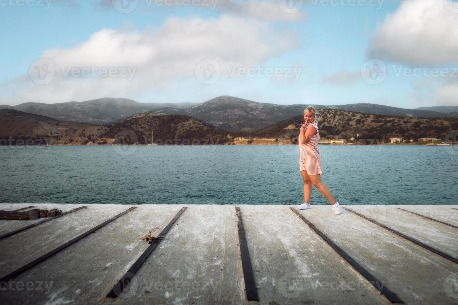 blonde vrouw in rode strepen witte jurk staande op het scheepsdok bij zonsondergang avond, glimlachend en kijkend naar de camera. toeristisch concept. argostoli, kefalonia, griekenland. foto