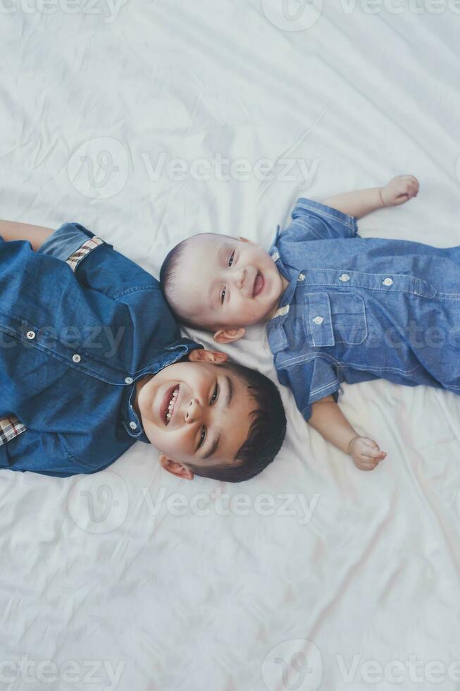 gelukkig kinderjaren concept. gelukkig broers portret. 6 jaren jongen en 6 maanden oud baby jongen hebben plezier. twee weinig kinderen glimlachen naar de camera. schattig weinig broers aan het liegen Aan bed samen foto