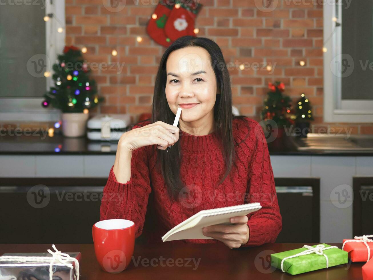 Aziatisch vrouw vervelend rood gebreid trui zittend Bij tafel met rood kop van koffie en geschenk dozen in de keuken met Kerstmis decoratie, gebruik makend van pen schrijven naam lijst Aan notebook. foto