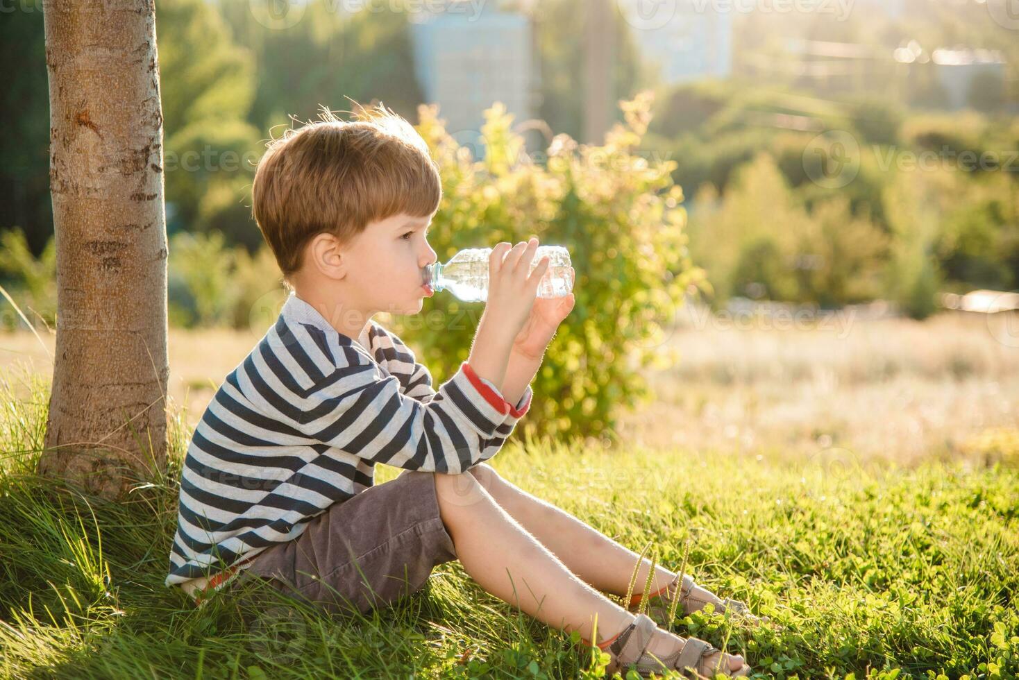 schattig jongen zittend Aan de gras drankjes water van een fles in de zomer Bij zonsondergang. kind blust dorst Aan een heet dag foto