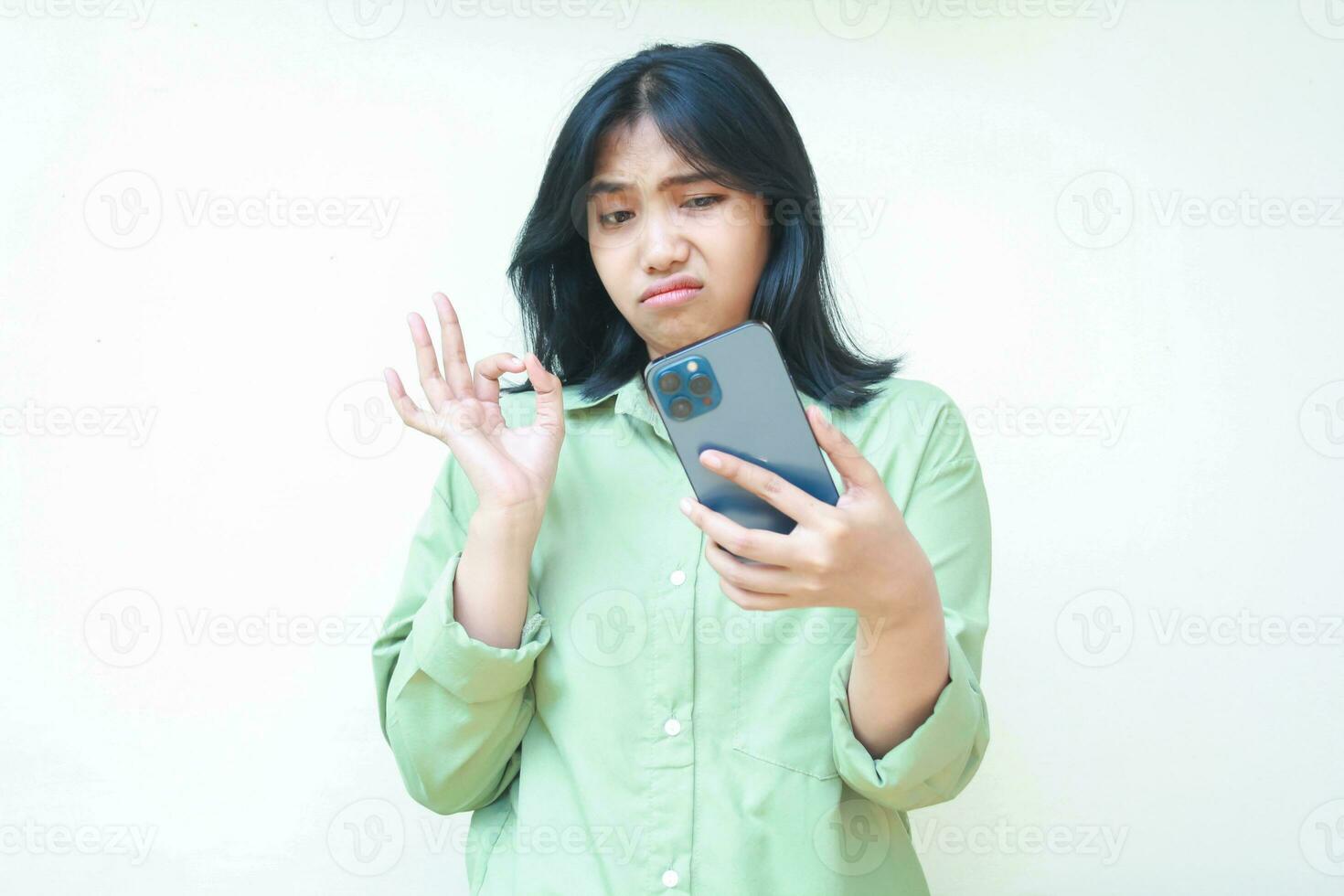 onwetend aarzelend Aziatisch vrouw vervelend over- formaat groen overhemd tonen OK tekens met twijfelen ongeloof uitdrukking terwijl vergadering online via smartphone geïsoleerd Aan wit achtergrond foto