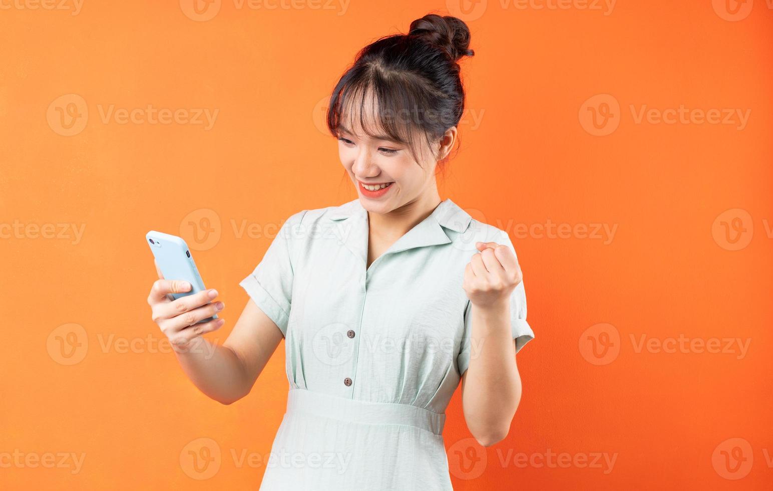Portret van een jong meisje dat telefoon gebruikt en de overwinning viert, geïsoleerd op een oranje achtergrond foto