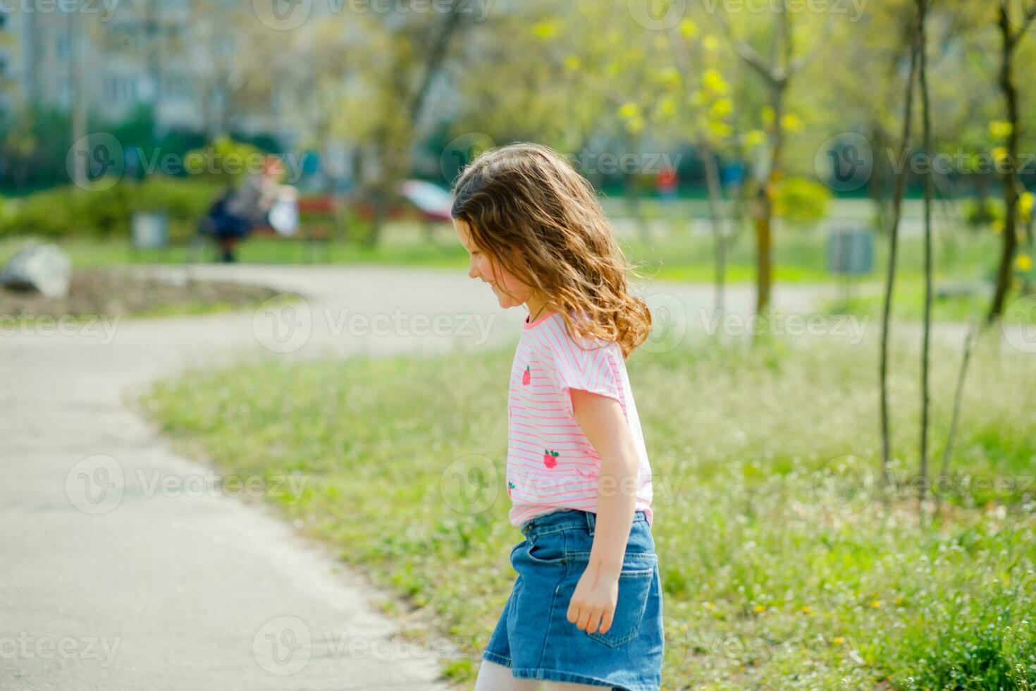 mooi weinig meisje met ontwikkelen haar- wandelingen in straat schenden quarantaine van coronavirus. meisje in roze t-shirt loopt aan de overkant groen veld. meisje Toneelstukken actief spellen in natuur in zomer of de lente. foto