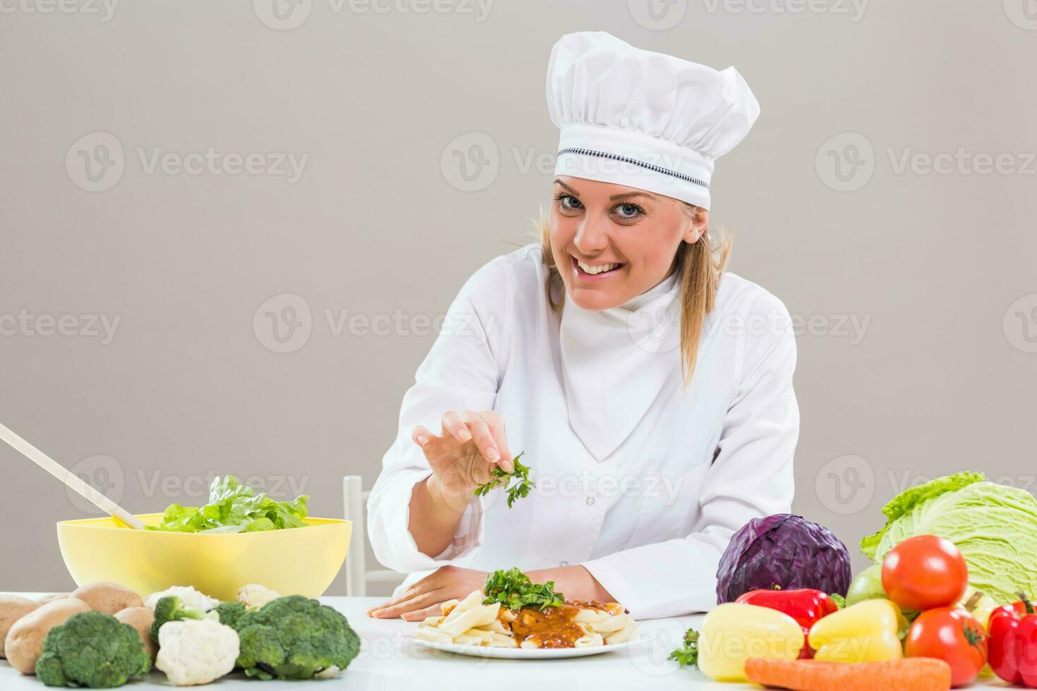 vrolijk vrouw chef is zittend Bij de tafel met bundel van groente en decoreren bereid maaltijd. foto