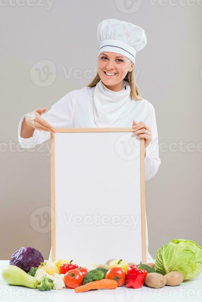 vrolijk vrouw chef is tonen leeg whiteboard met bundel van groenten Aan de tafel. foto