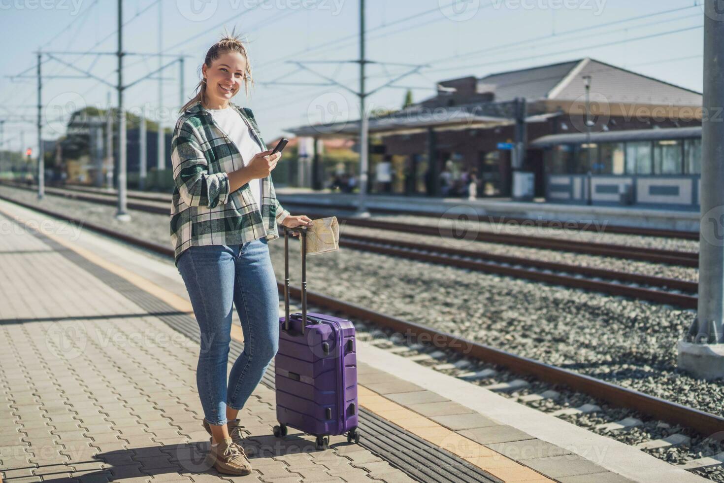 gelukkig vrouw gebruik makend van mobiel telefoon en Holding kaart Aan een trein station foto