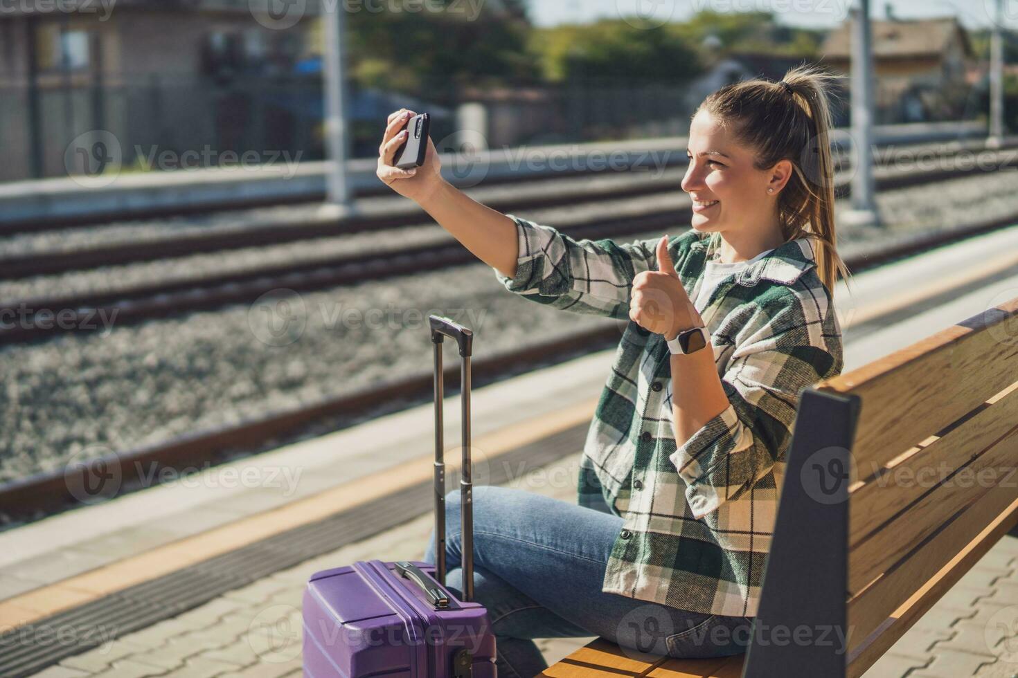 gelukkig vrouw tonen duim omhoog terwijl nemen selfie met mobiel telefoon Aan een trein station foto