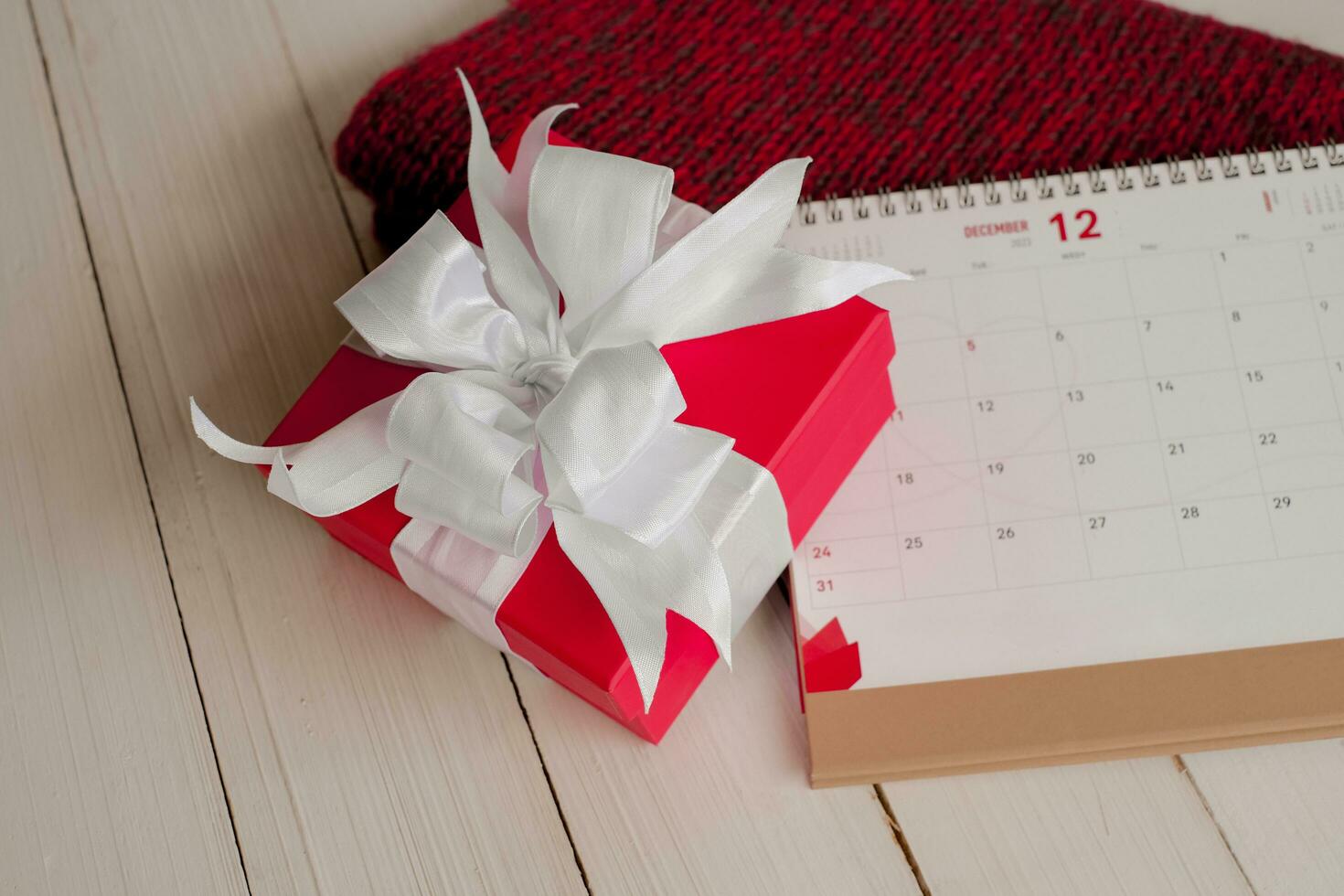 rood geschenk doos en kalender Aan geïsoleerd Aan een rood gebreid sjaal met wit houten achtergrond met. kerstmis dag concept. planning het roosteren agenda, evenement, organisator Kerstmis dag. vlak leggen, top visie. foto