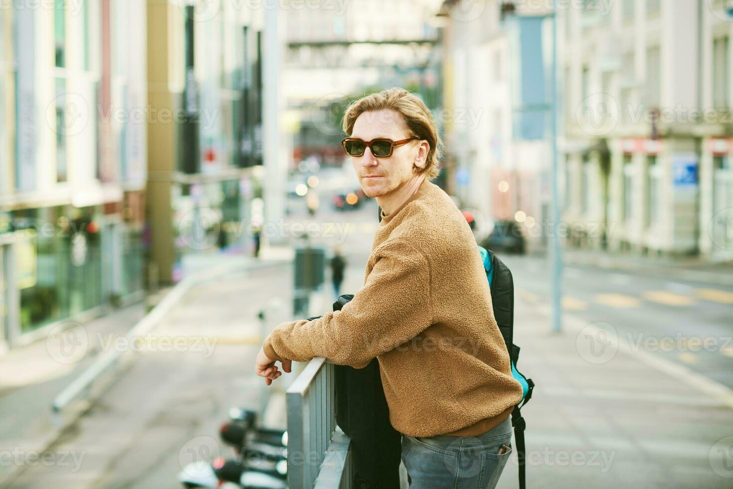 buitenshuis portret van knap jong Mens vervelend zonnebril en beige wazig fleece trui, poseren Aan stad straat achtergrond foto