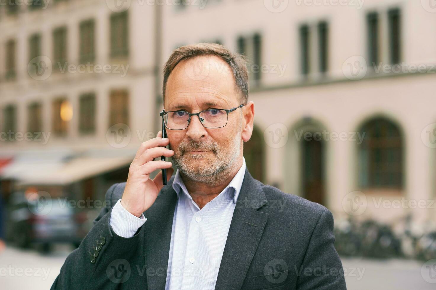 buitenshuis portret van midden- leeftijd Mens, vervelend bril en grijs pak, Holding smartphone foto