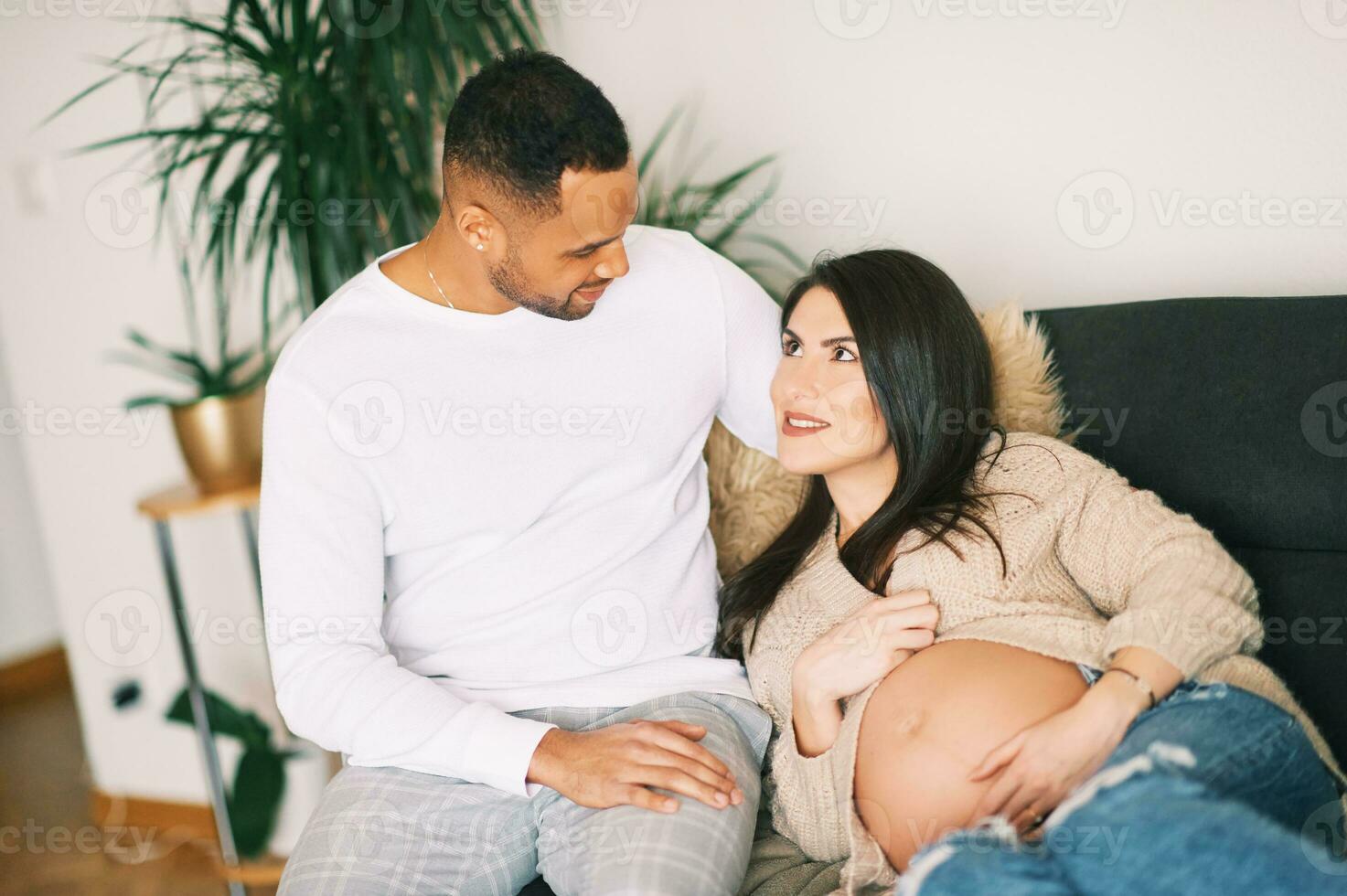 binnen- portret van gelukkig jong stel, zwanger vrouw resting Aan een bankstel met haar liefhebbend man foto
